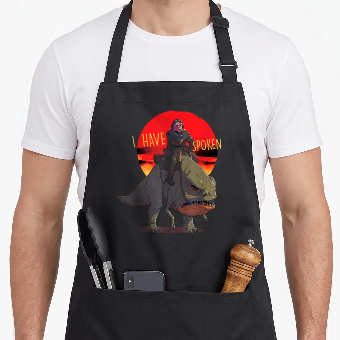 Kuiil spokane apron - I HAVE SPOKEN text flanking a person riding a dinosaur before a red sun — shown on a black cotton canvas apron against a white background