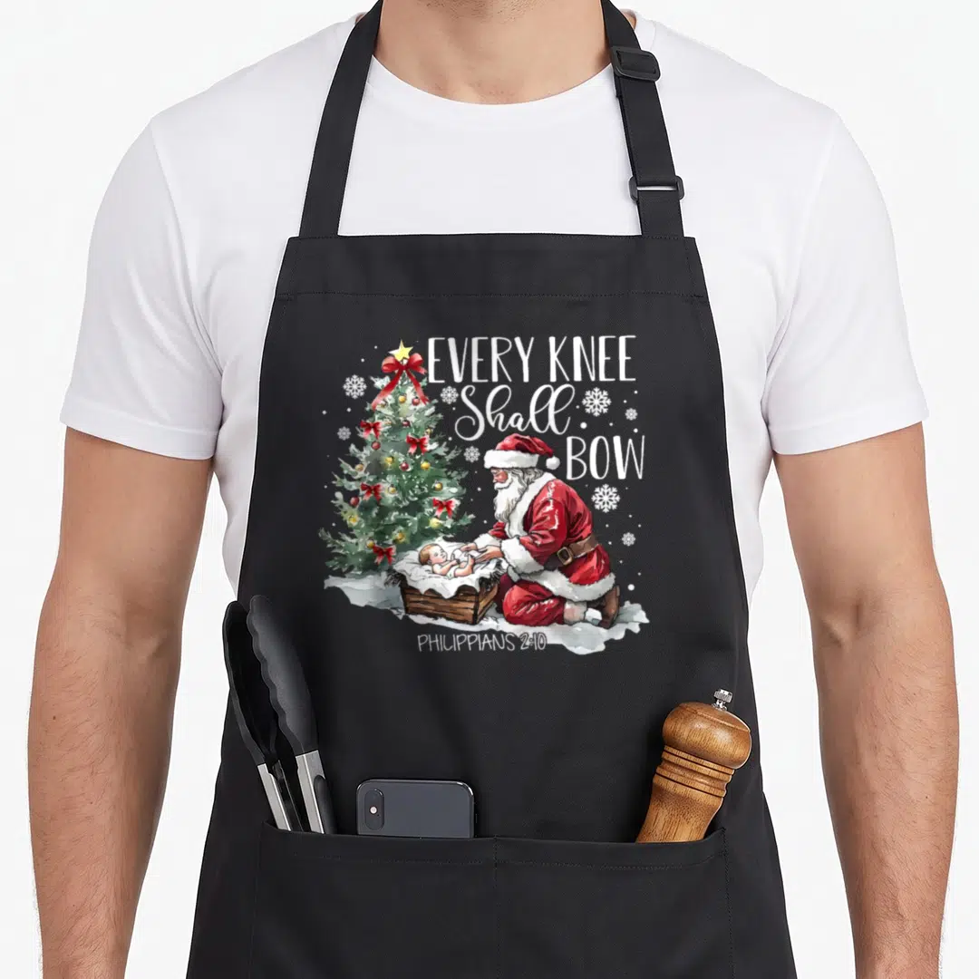 Knee christmas santa apron - Santa kneeling at a manger beside a Christmas tree with Every Knee Shall Bow text — shown on a black cotton canvas apron against a white background