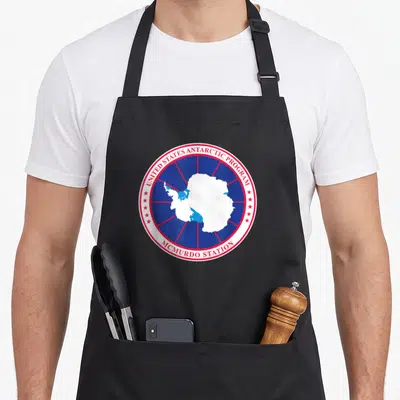 Arctic traveler apron - Circular emblem with white Antarctica map, red stars, and McMurdo Station text on blue — shown on a black cotton canvas apron against a white background