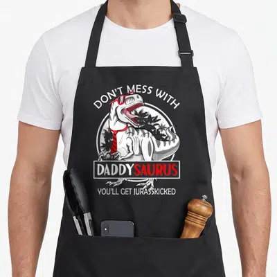 Daddysaurus mess apron - T-Rex wearing red glasses and tie with pun text about not messing with daddy — shown on a black cotton canvas apron against a white background
