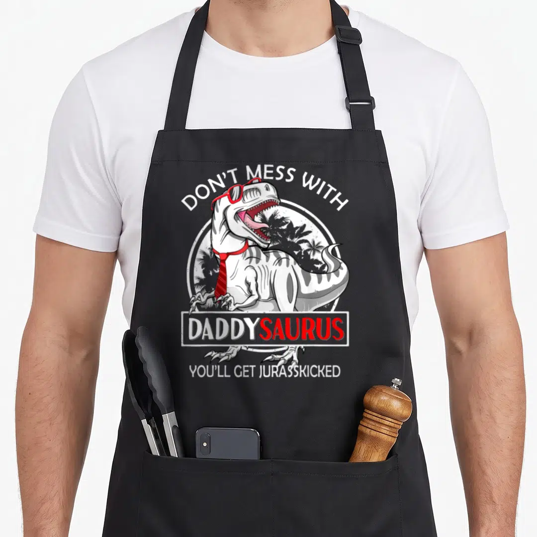 Daddysaurus mess apron - T-Rex wearing red glasses and tie with pun text about not messing with daddy — shown on a black cotton canvas apron against a white background