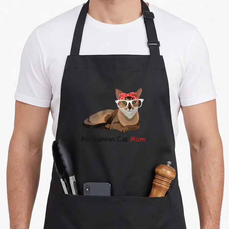 Abyssinian cat apron - Cartoon Abyssinian cat wearing white glasses and red bandana above text — shown on a black cotton canvas apron against a white background