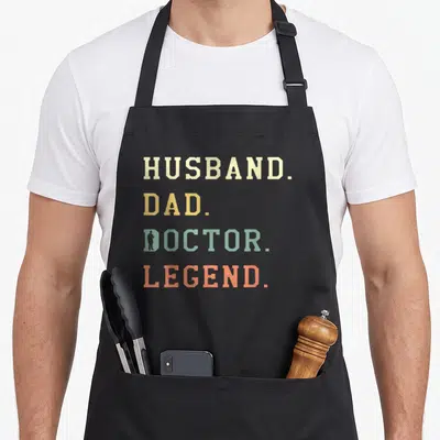Legend doctor with apron - Stacked text HUSBAND DAD DOCTOR LEGEND in cream yellow teal orange with silhouette inside D — shown on a black cotton canvas apron against a white background