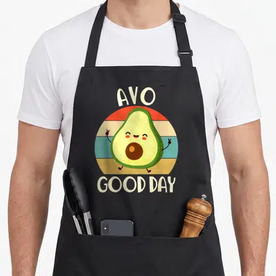 Toddler avocado apron - Cartoon avocado with peace signs inside striped circle, AVO above, GOOD DAY below — shown on a black cotton canvas apron against a white background
