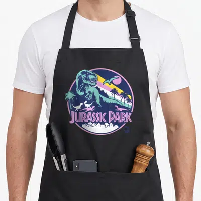 Scene dinosaur apron - Retro circular T-Rex and Pterosaur design with JURASSIC PARK text and palm trees in blue pink — shown on a black cotton canvas apron against a white background