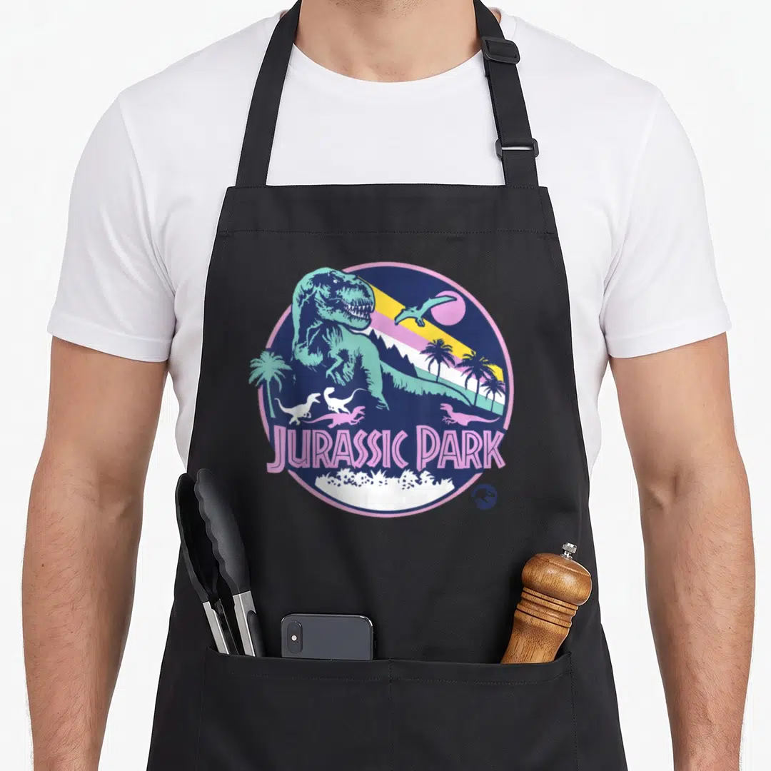 Scene dinosaur apron - Retro circular T-Rex and Pterosaur design with JURASSIC PARK text and palm trees in blue pink — shown on a black cotton canvas apron against a white background
