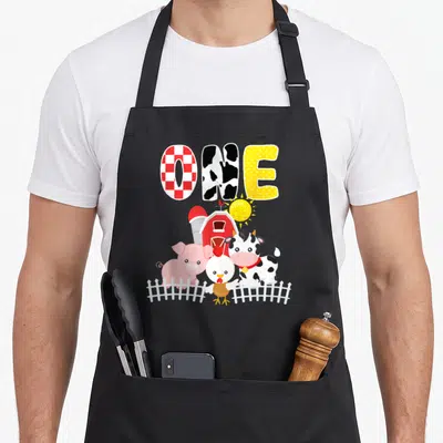 Animal 1st farm aprons - Cartoon cow pig chicken behind white fence with patterned ONE text — shown on a black cotton canvas apron against a white background