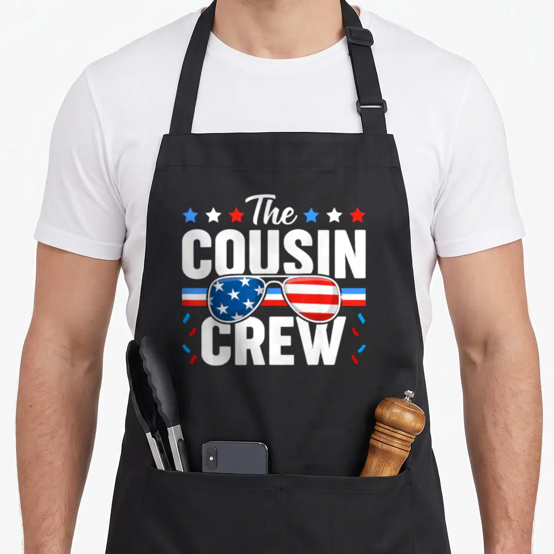 Cousin 4th of july apron - Sunglasses with US flag patterns and stars flanking The Cousin Crew text in white, red — shown on a black cotton canvas apron against a white background