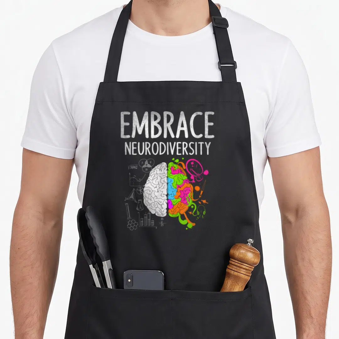 April teacher art apron - EMBRACE NEURODIVERSITY text above split brain with white science symbols and multicolored art — shown on a black cotton canvas apron against a white background