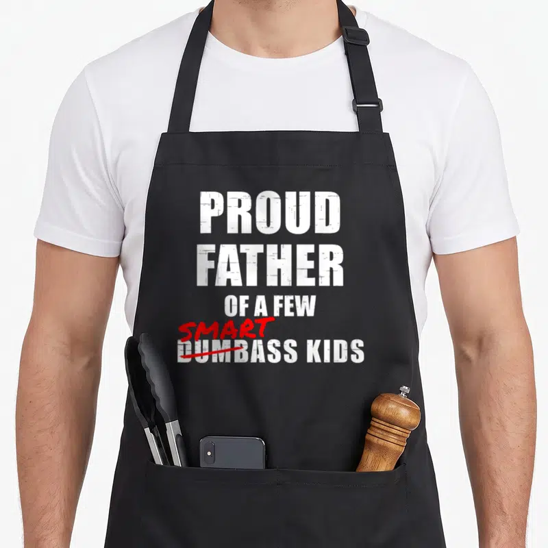 Father proud dad apron - Stacked white text reading Proud Father Of A Few Smart Dumbass Kids with red cross-out — shown on a black cotton canvas apron against a white background