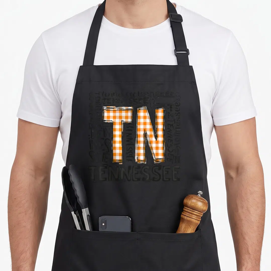 Tennessee the plaid apron - Central TN monogram with orange white plaid fill surrounded by stacked Tennessee text — shown on a black cotton canvas apron against a white background