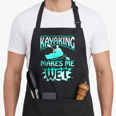 Watersport wet aprons - Cartoon kayaker silhouette with KAYAKING MAKES ME WET text in turquoise and white splash shape — shown on a black cotton canvas apron against a white background