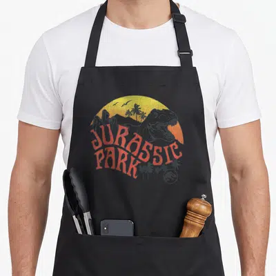 Sunset t-rex apron - Grey T-Rex head over yellow orange sun with red Jurassic Park text and palm trees — shown on a black cotton canvas apron against a white background
