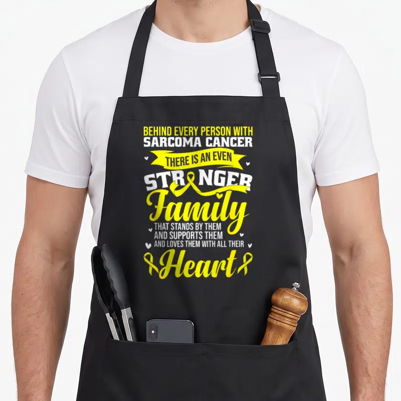 Sarcoma cancer awareness ribbon apron - Yellow and white stacked text reading Behind Every Person With Sarcoma Cancer — shown on a black cotton canvas apron against a white background