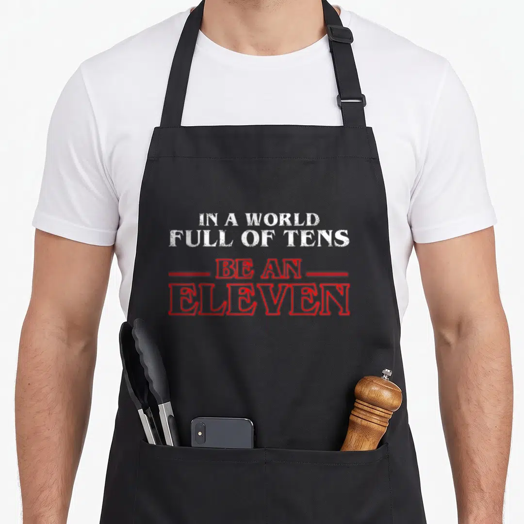 Eleven novelty apron - Stacked white and red text reading IN A WORLD FULL OF TENS BE AN ELEVEN — shown on a black cotton canvas apron against a white background