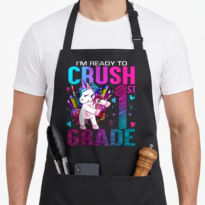 Crush 1st unicorn apron - Cartoon unicorn with pencils and I'M READY TO CRUSH 1ST GRADE text in pink blue purple — shown on a black cotton canvas apron against a white background