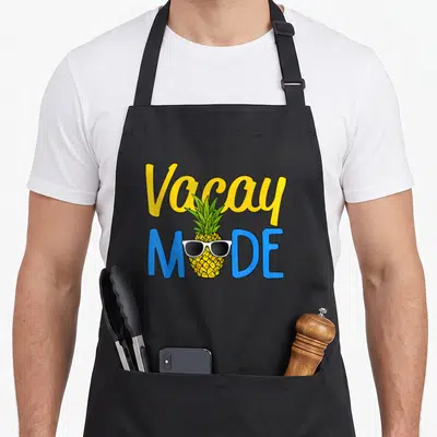 Vacay holiday apron - Cartoon pineapple in sunglasses between yellow Vacay and blue MODE text — shown on a black cotton canvas apron against a white background