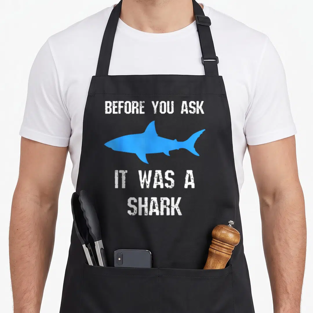 Amputation amputee awareness apron - Blue shark silhouette between white distressed text reading Before You Ask It Was — shown on a black cotton canvas apron against a white background