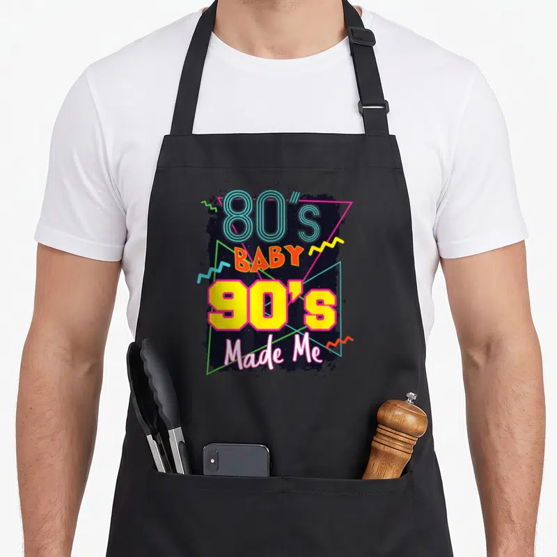 Baby 80s apron - Stacked retro text 80s baby 90s made me with geometric triangles and zigzags on dark splatter — shown on a black cotton canvas apron against a white background