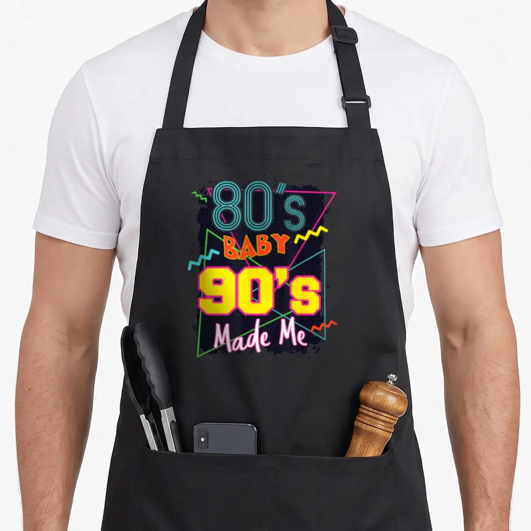 Baby 80s apron - Stacked retro text 80s baby 90s made me with geometric triangles and zigzags on dark splatter — shown on a black cotton canvas apron against a white background