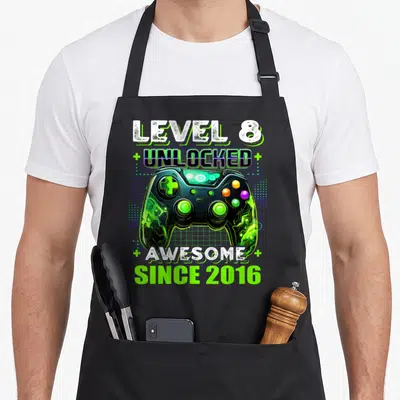 8th gamer birthday apron - Game controller with Level 8 Unlocked Awesome Since 2016 text and green plus signs on blue — shown on a black cotton canvas apron against a white background