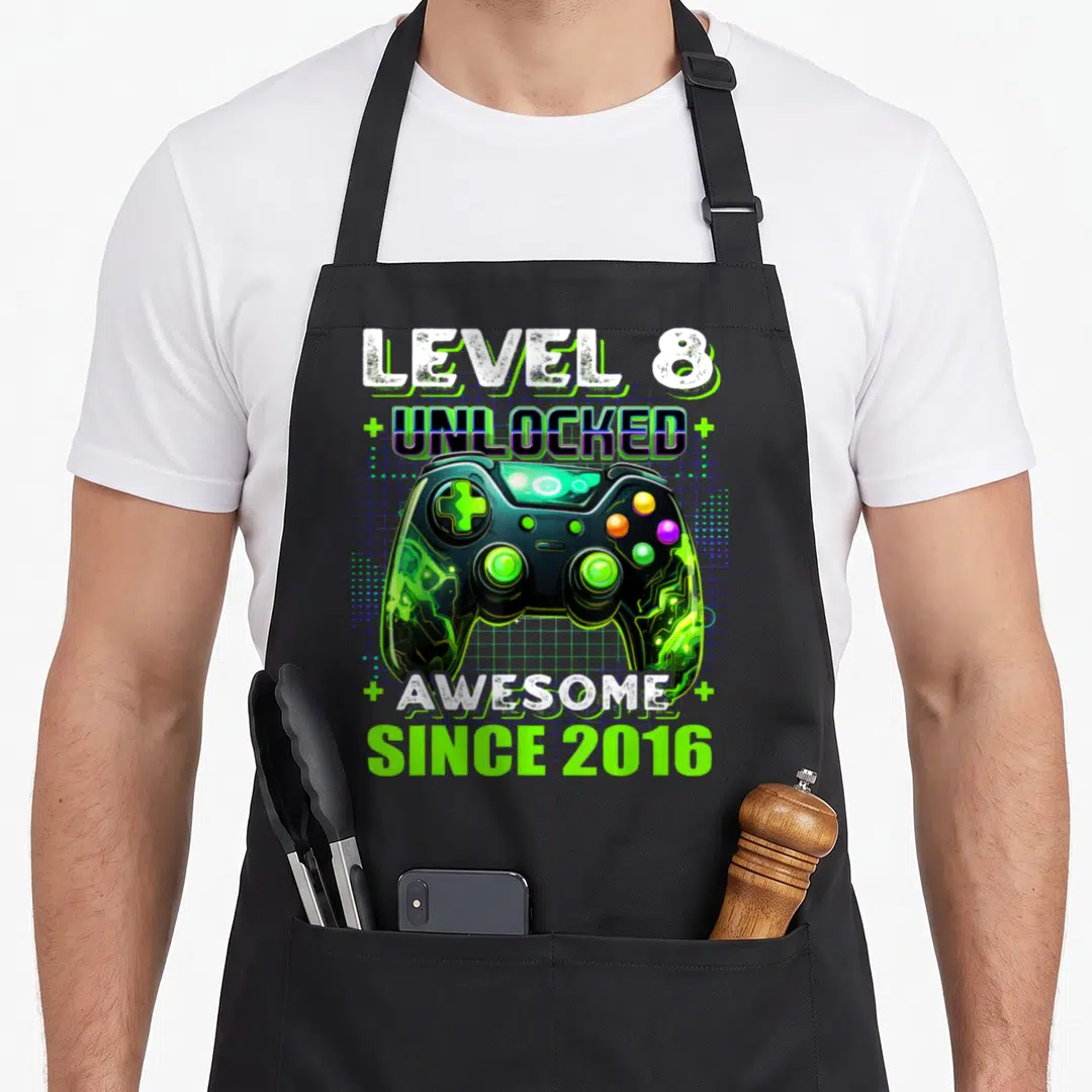 8th gamer birthday apron - Game controller with Level 8 Unlocked Awesome Since 2016 text and green plus signs on blue — shown on a black cotton canvas apron against a white background