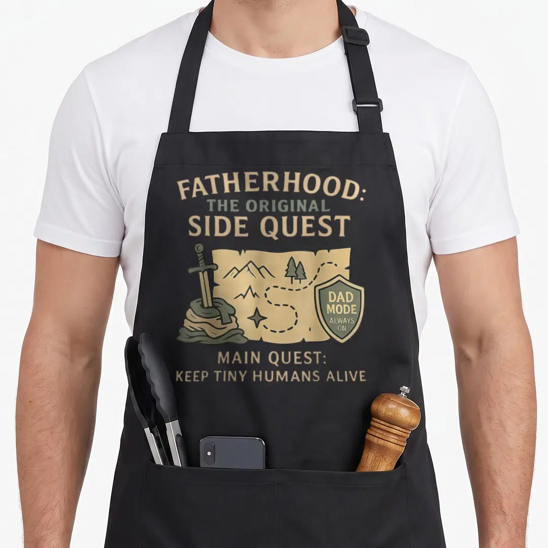 Father tiny apron - Map graphic with sword in rock, shield reading Dad Mode Always — shown on a black cotton canvas apron against a white background