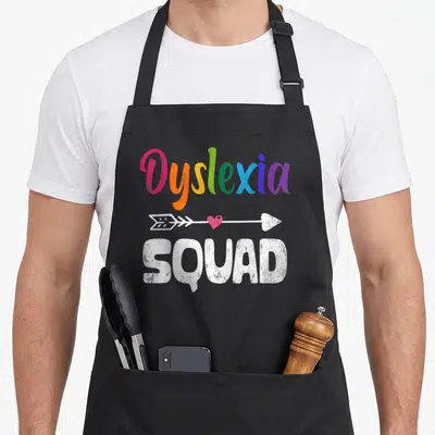 Awareness teacher apron - Rainbow Dyslexia text above white arrow with pink heart and SQUAD below — shown on a black cotton canvas apron against a white background