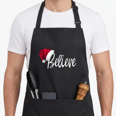 Christmas believe santa claus apron - White Believe script with red Santa hat perched on letter B and star above i — shown on a black cotton canvas apron against a white background