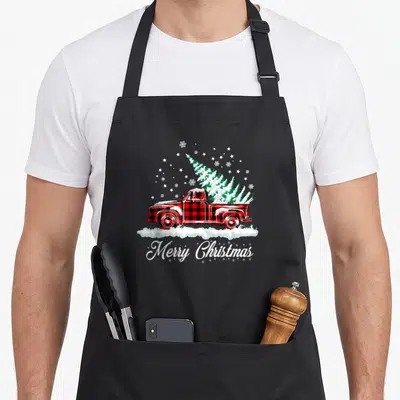 Plaid christmas tree apron - Red plaid truck carrying snow covered green tree with Merry Christmas text below — shown on a black cotton canvas apron against a white background