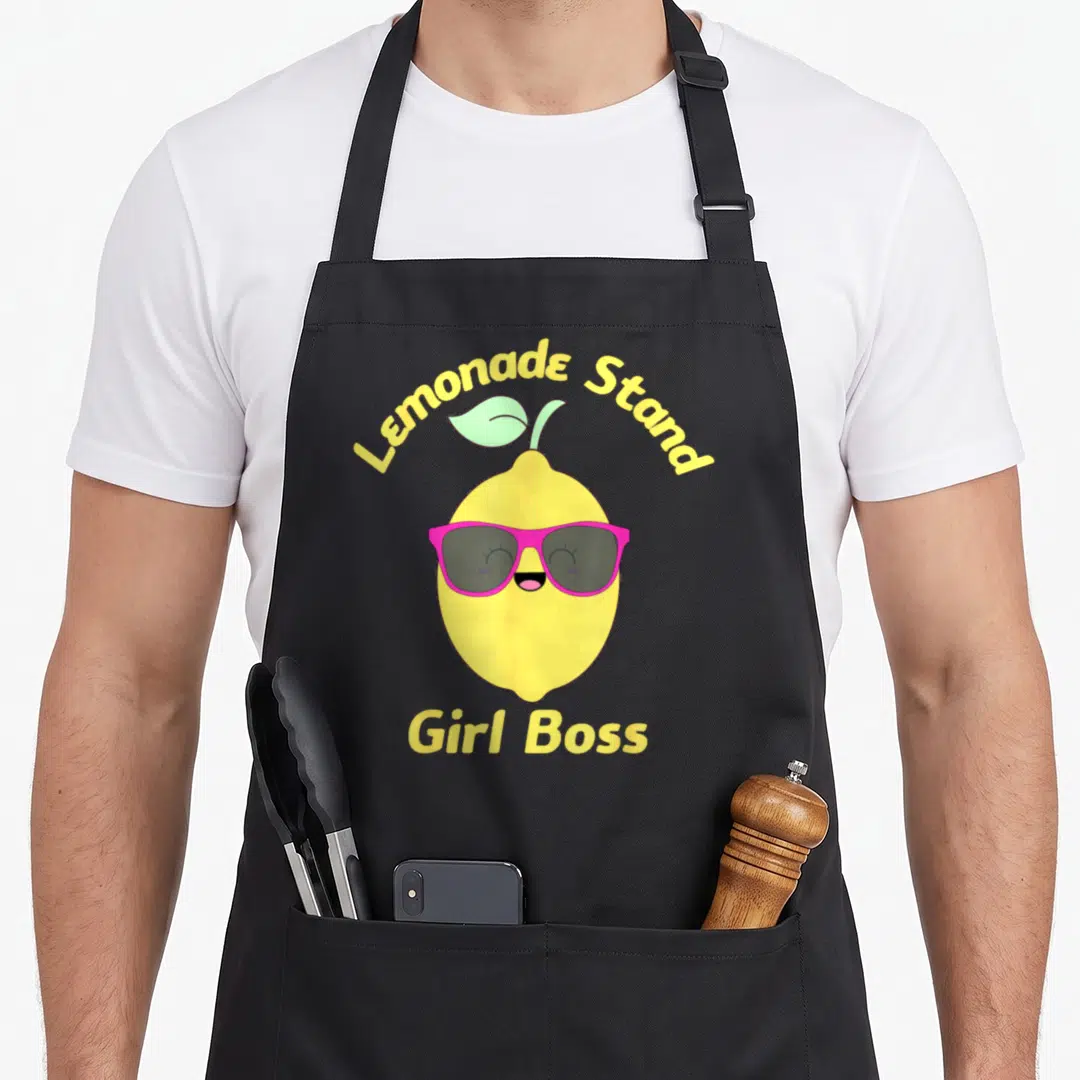 Fruit lemonade apron - Cartoon lemon wearing pink sunglasses with Lemonade Stand Girl Boss text — shown on a black cotton canvas apron against a white background