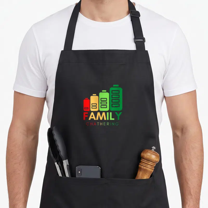 Cute family gathering apron - Four red yellow and green batteries above FAMILY CHATHERING text — shown on a black cotton canvas apron against a white background
