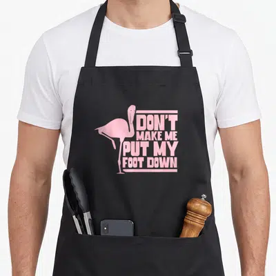 Foot animal flamingo apron - Pink flamingo silhouette standing on one leg beside stacked text 'DON'T MAKE ME PUT MY — shown on a black cotton canvas apron against a white background
