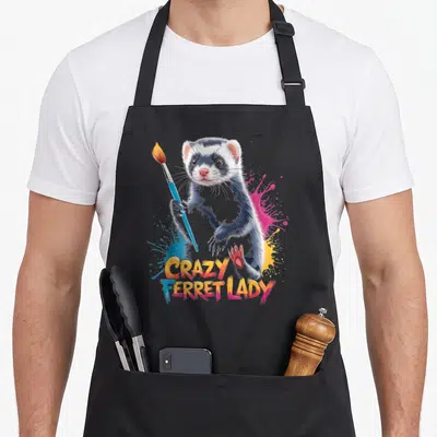 Ferret colorful aprons - Realistic ferret holding paintbrush with colorful splatters and gradient text reading CRAZY — shown on a black cotton canvas apron against a white background