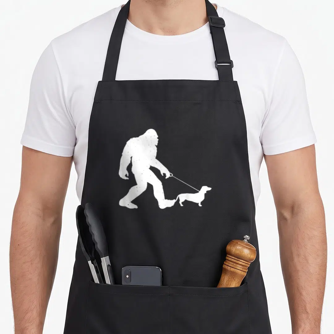 Bigfoot wiener apron - White distressed silhouette of Bigfoot walking a dachshund on a leash — shown on a black cotton canvas apron against a white background