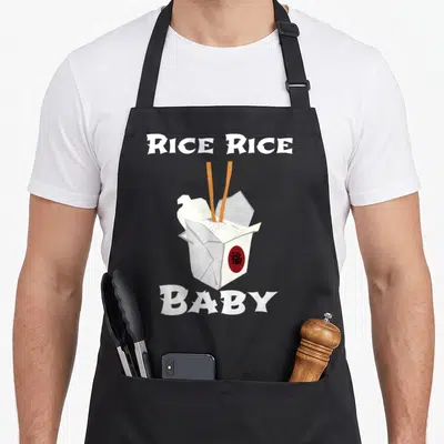 Baby asian apron - RICE RICE BABY text above and below cartoon takeout box with orange chopsticks and red seal — shown on a black cotton canvas apron against a white background