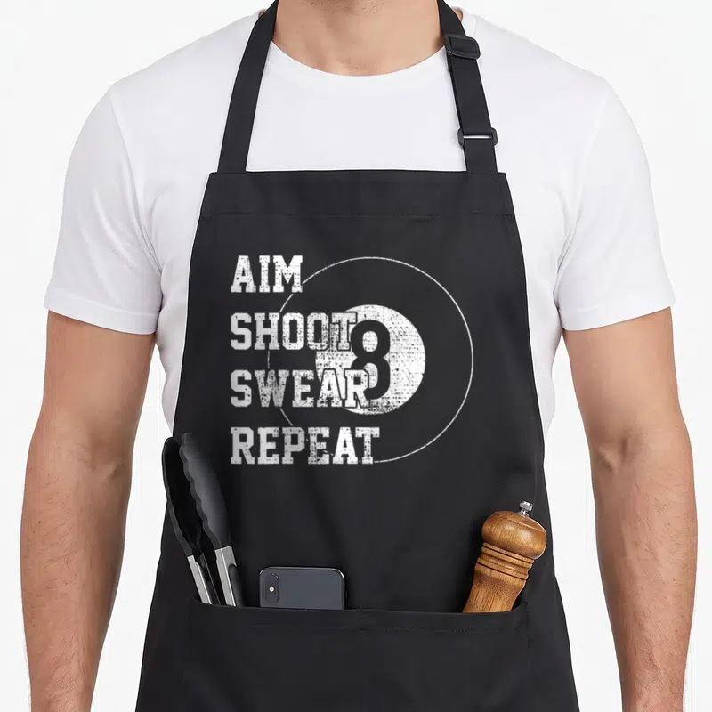Ball billiards lover apron - White distressed AIM SHOOT SWEAR REPEAT text next to a large 8-ball inside a thin circle — shown on a black cotton canvas apron against a white background