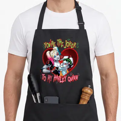 Cute vehicle apron - Harley Quinn and Joker inside red heart with YOU'RE THE JOKER TO MY HARLEY QUINN text in red green — shown on a black cotton canvas apron against a white background