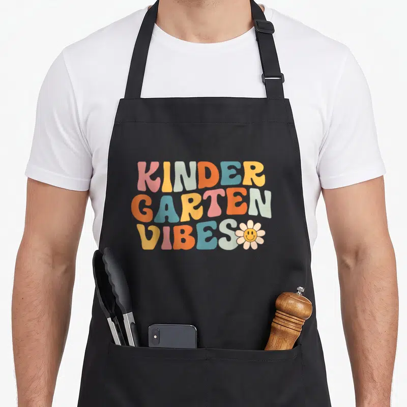 Kindergarten crew apron - Stacked KNDER GARTEN VIBES text in pink yellow blue orange grey with small smiley flower — shown on a black cotton canvas apron against a white background