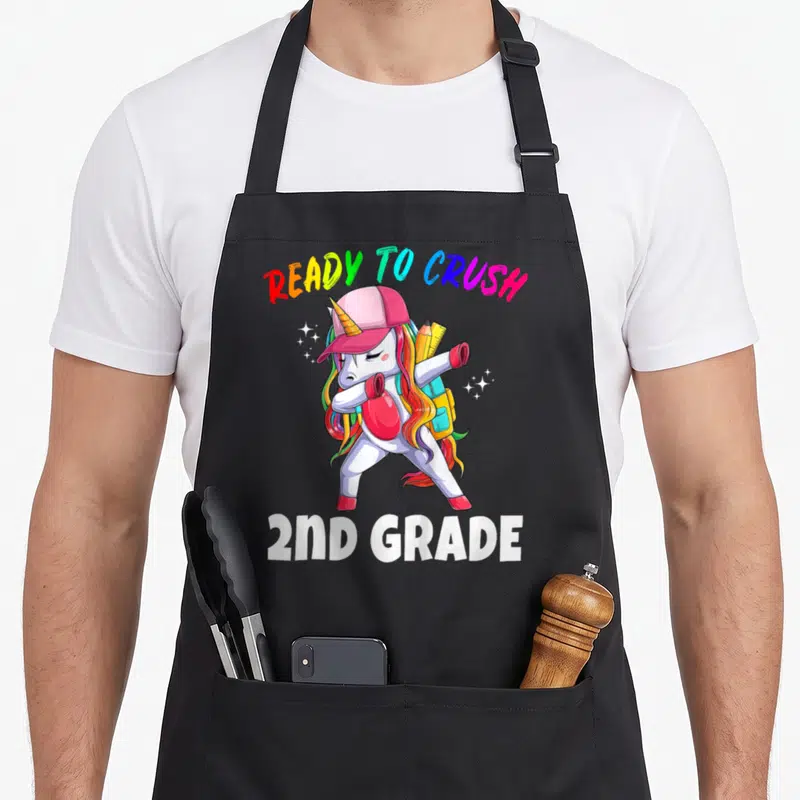 2nd grade unicorn apron - Dabbing unicorn with pink cap, backpack, and boxing glove under rainbow text — shown on a black cotton canvas apron against a white background