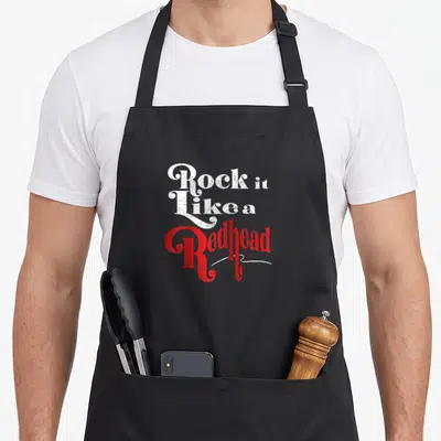 Redhead baseball apron - Stacked distressed white and red text reading Rock It Like a Redhead on black fabric — shown on a black cotton canvas apron against a white background
