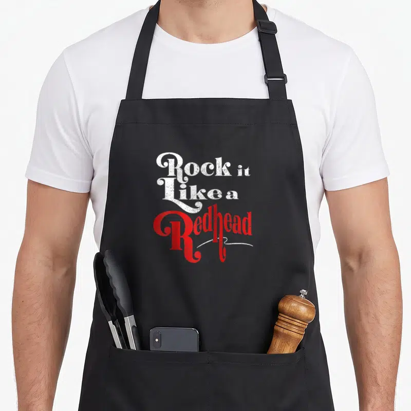 Redhead baseball apron - Stacked distressed white and red text reading Rock It Like a Redhead on black fabric — shown on a black cotton canvas apron against a white background