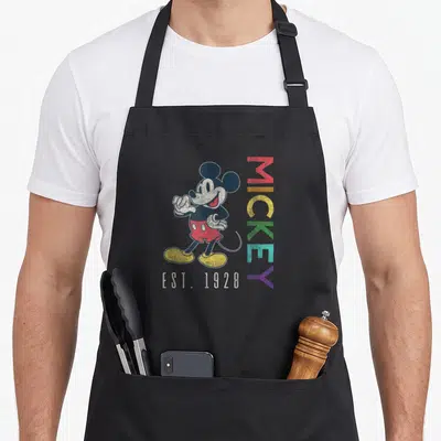 Est rainbow apron - Mickey Mouse standing next to vertical rainbow MICKEY text and EST. 1928 below — shown on a black cotton canvas apron against a white background