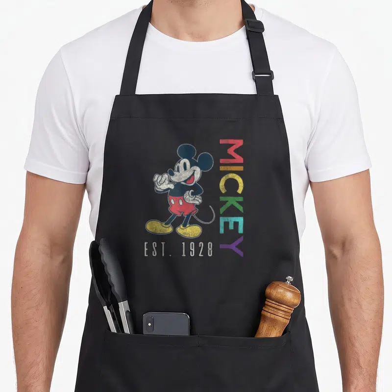 Est rainbow apron - Mickey Mouse standing next to vertical rainbow MICKEY text and EST. 1928 below — shown on a black cotton canvas apron against a white background
