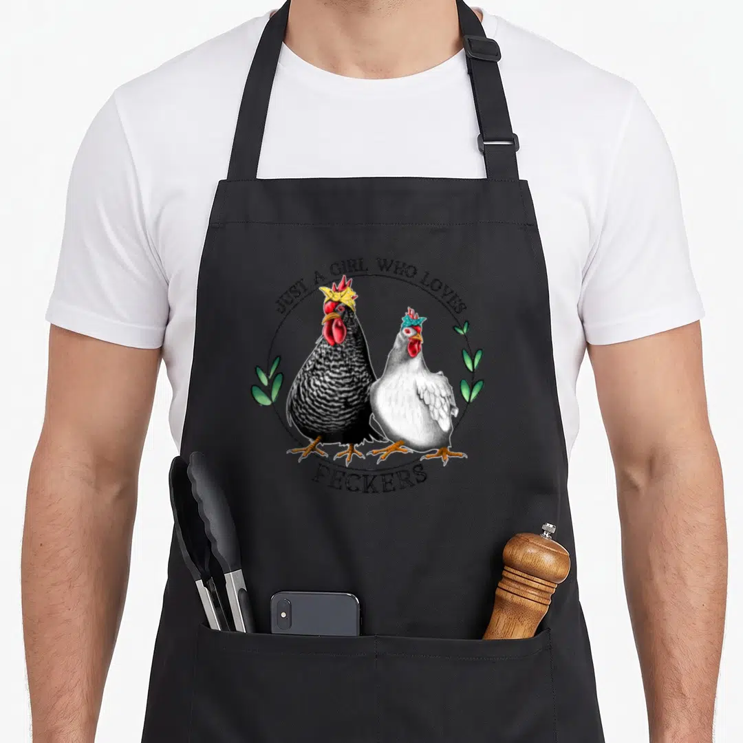 Pecker chicken apron - Two crowned hens in black circle with green leaves and peckers text — shown on a black cotton canvas apron against a white background