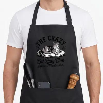Crazy cat woman apron - Line art of a tabby cat on a cushion with THE CRAZY Cat Lady Club Lifetime Membership text — shown on a black cotton canvas apron against a white background