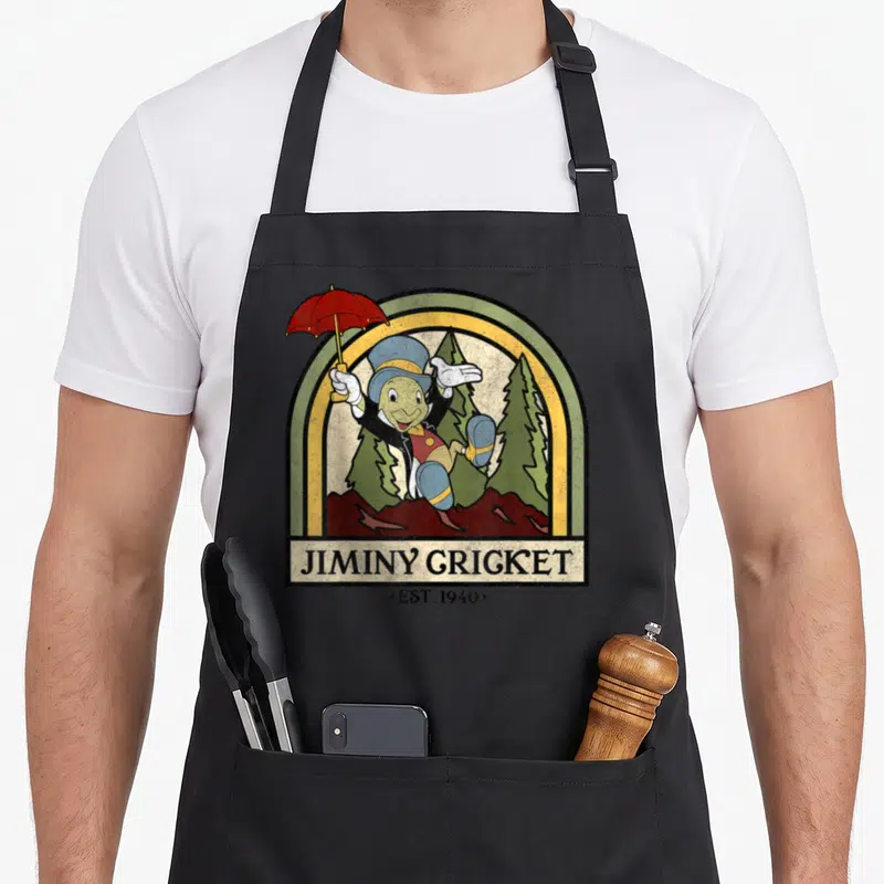 Pinocchio cricket apron - Jiminy Cricket cartoon with red umbrella in rainbow arch, EST. 1940 text below — shown on a black cotton canvas apron against a white background