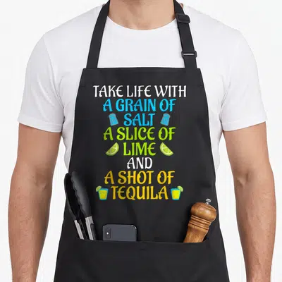 Salt agave apron - Stacked text take life with salt lime tequila flanked by shakers limes and glasses — shown on a black cotton canvas apron against a white background