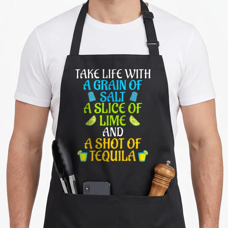 Salt agave apron - Stacked text take life with salt lime tequila flanked by shakers limes and glasses — shown on a black cotton canvas apron against a white background