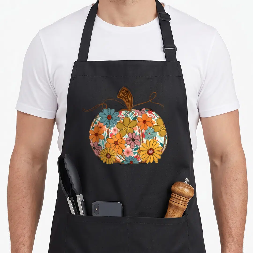 Floral fall aprons - Cartoon pumpkin filled with orange blue yellow pink flowers and brown vines — shown on a black cotton canvas apron against a white background